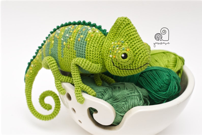 Puede incluir: Un juguete de camale&oacute;n de crochet verde con manchas amarillas, sentado en un cuenco de lana blanco con ovillos de lana verdes. El juguete tiene una cara sonriente y una cola enroscada.