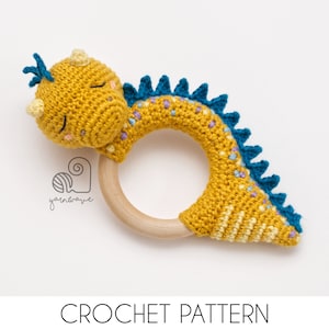 May include: A crocheted yellow dinosaur rattle with a blue and purple spiky tail, attached to a wooden ring. The rattle is a handmade baby toy.
