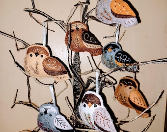 May include: A collection of handmade felt owl ornaments in various colors, including tan, brown, gray, and blue. Each owl has unique embroidered details and is suspended by a white ribbon. The ornaments are displayed on a decorative branch.