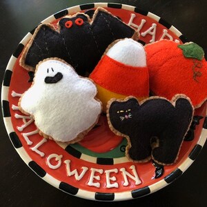 May include: A plate with a Halloween theme features felt toys shaped like a bat, ghost, candy corn, pumpkin, and black cat. The plate has a red background with black and white stripes and the words "Happy Halloween" written in white.