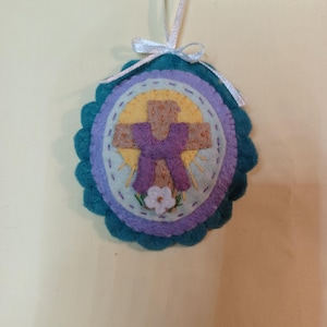 May include: A handmade felt ornament with a cross design. The ornament features a purple cross on a yellow background, surrounded by blue, purple, and teal felt layers. A white ribbon is attached for hanging. A small white flower is at the bottom of the cross.