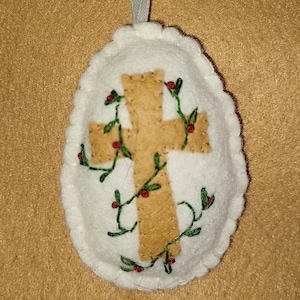 May include: White felt Easter egg ornament with a brown felt cross and green embroidered vines with red berries.