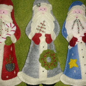 May include: Three felt Christmas ornaments, each depicting a Santa Claus figure. The Santa on the left is wearing a red suit with white trim and holding a candy cane. The middle Santa is wearing a gray suit with white trim and holding a wreath. The Santa on the right is wearing a blue suit with white trim and holding a star.
