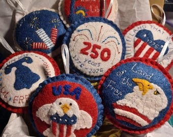 May include: A collection of patriotic felt ornaments in red, white, and blue. Designs include fireworks, the Statue of Liberty, an eagle, and the American flag. Some ornaments feature text like "USA" and "Land of the Free."