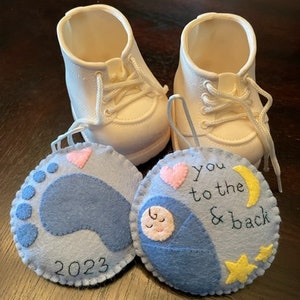 May include: Two felt ornaments, one with a blue footprint and the year 2023, the other with a sleeping baby and the text "you to the & back".