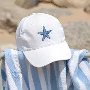 K&ouml;nnte beinhalten: Eine wei&szlig;e Baseballkappe mit einem blauen, gestickten Seestern-Design. Die Kappe liegt auf einem blau-wei&szlig; gestreiften Handtuch, was auf ein Strand- oder Sommerthema hindeutet. Die Kappe besteht aus einem glatten Material.