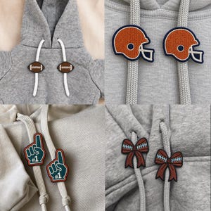 May include: A gray hooded sweatshirt featuring white drawstrings with various embroidered attachments. These include brown footballs, orange football helmets, green foam fingers with the text #1, and brown bows with white stitching details.