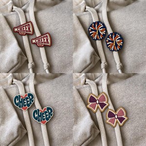 May include: A collection of four pairs of embroidered patches attached to light beige hoodie drawstrings. The designs include red megaphone patches with the word CHEER, blue and orange pom-pom patches, heart-shaped patches with the word CHEER, and pink and purple bow patches.