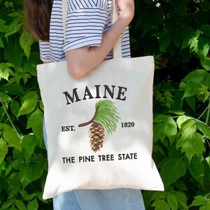 May include: A cream tote bag featuring the word "MAINE" embroidered in black. Below is a detailed illustration of a pine cone and needles. The text "EST. 1820" and "THE PINE TREE STATE" are also printed on the bag.
