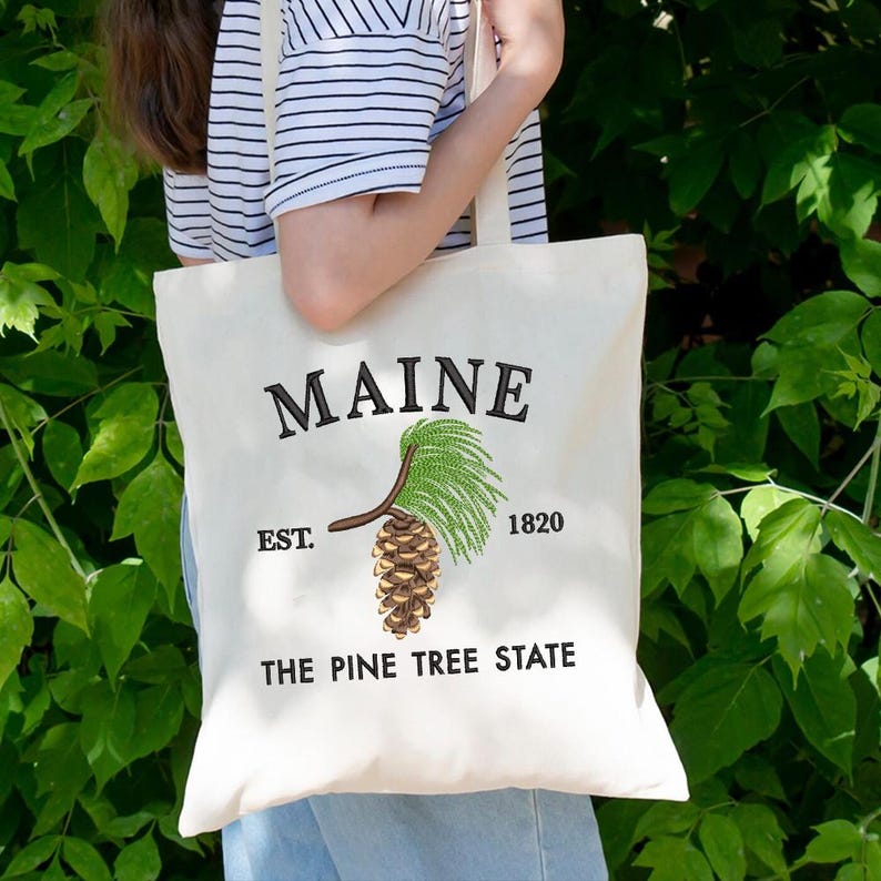 May include: A cream tote bag featuring the word "MAINE" embroidered in black. Below is a detailed illustration of a pine cone and needles. The text "EST. 1820" and "THE PINE TREE STATE" are also printed on the bag.