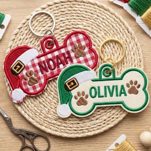May include: Two bone-shaped keychains with metal rings. One is red and white gingham with a Santa hat and the name NOAH embroidered in red. The other is cream with a green Santa hat and the name OLIVIA embroidered in green. Both feature brown paw prints.