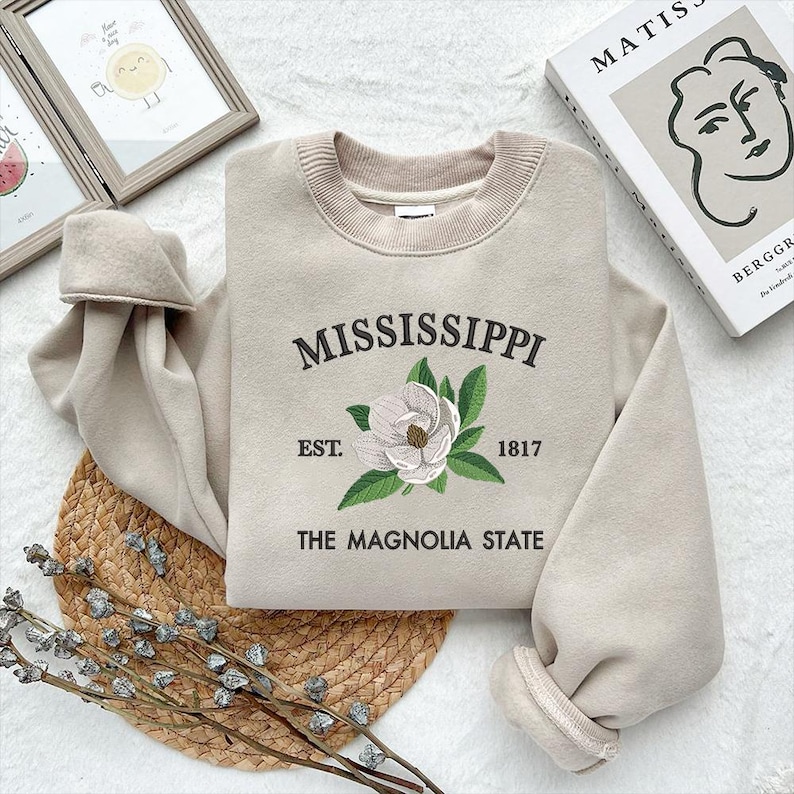 May include: Beige sweatshirt featuring the embroidered word "MISSISSIPPI", a magnolia flower, and the text "EST. 1817 THE MAGNOLIA STATE". It has a crew neck and long sleeves.