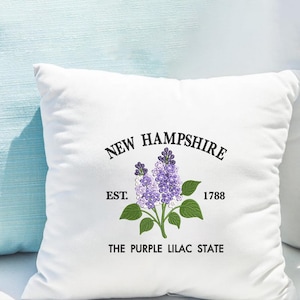May include: White square cushion featuring an embroidered design. The design includes the text "NEW HAMPSHIRE" above a purple lilac flower with green leaves. The text "EST. 1788" and "THE PURPLE LILAC STATE" are also included.