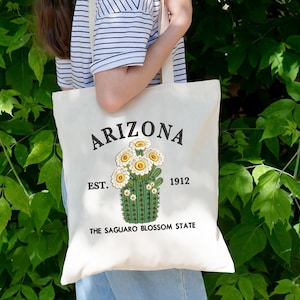 May include: Cream tote bag featuring the word "ARIZONA" embroidered in black, with a cactus and flower design. The text "EST. 1912" and "THE SAGUARO BLOSSOM STATE" are also present. The bag has a simple, practical design.