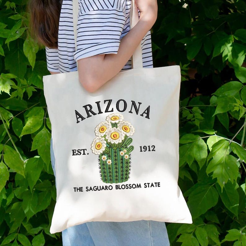 May include: Cream tote bag featuring the word "ARIZONA" embroidered in black, with a cactus and flower design. The text "EST. 1912" and "THE SAGUARO BLOSSOM STATE" are also present. The bag has a simple, practical design.