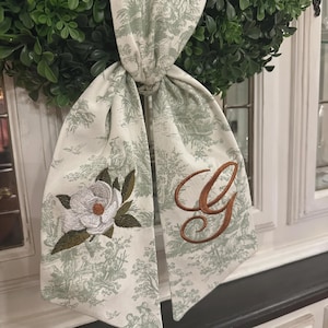 May include: A decorative fabric bow featuring a sage green and white toile de Jouy pattern. The left tail displays an embroidered white magnolia flower with green leaves, while the right tail features a large, brown embroidered letter G monogram.