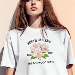 May include: White t-shirt featuring a floral design with the text "NORTH CAROLINA" above a floral illustration and the text "EST. 1789 THE DOGWOOD STATE". The design includes pink flowers and green leaves.