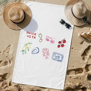 May include: White beach towel with colorful illustrations of olives, lemons, tomatoes, sunglasses, an oyster, a lobster, and a sardine can. The words "Dolce Vita" are printed in red. Two straw hats, sunglasses, and sandals are on the sand.