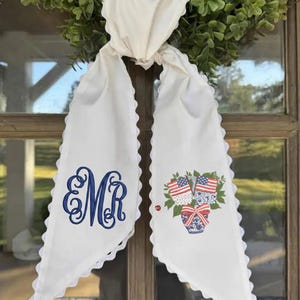 May include: White fabric door hanger with a scalloped edge. Features a blue embroidered monogram and a floral arrangement with American flags in a blue and white vase. The hanger is tied with a white ribbon.