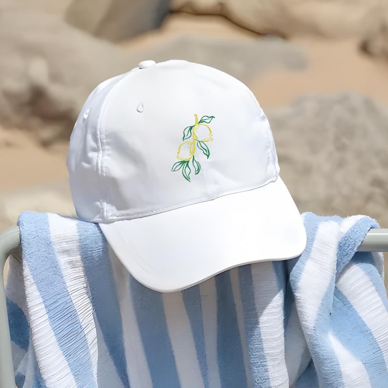 May include: A white baseball cap with a curved brim, featuring an embroidered lemon branch design in yellow and green. The cap rests on a blue and white striped towel, suggesting a beach or outdoor theme.