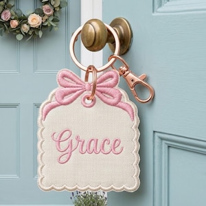 May include: A cream-colored fabric tag with scalloped edges and a pink embroidered bow at the top. The name Grace is embroidered in pink script in the center. The tag is attached to a rose gold metal keyring with a lobster clasp attachment.