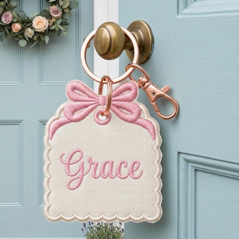 May include: A cream-colored fabric tag with scalloped edges and a pink embroidered bow at the top. The name Grace is embroidered in pink script in the center. The tag is attached to a rose gold metal keyring with a lobster clasp attachment.