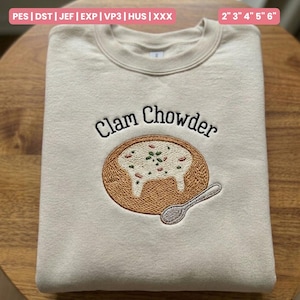 Puede incluir: Sudadera beige con un diseño bordado de sopa de almejas en un cuenco de pan, con una cuchara. El texto "Clam Chowder" está bordado encima del cuenco. La sudadera está doblada sobre una superficie de madera. Medidas: 5 cm, 7.6 cm, 10.2 cm, 12.7 cm, 15.2 cm.