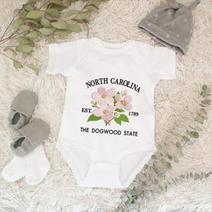 May include: White baby onesie with the text "NORTH CAROLINA" above a floral design and "EST. 1789 THE DOGWOOD STATE" below. Gray baby shoes, socks, and a gray knotted hat are also in the image.