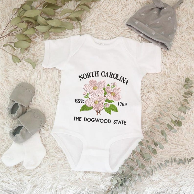May include: White baby onesie with the text "NORTH CAROLINA" above a floral design and "EST. 1789 THE DOGWOOD STATE" below. Gray baby shoes, socks, and a gray knotted hat are also in the image.