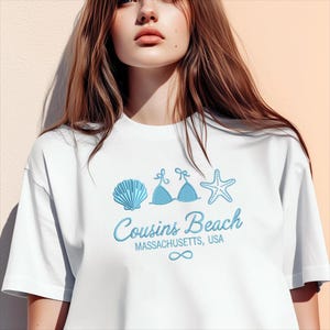 Puede incluir: Camiseta blanca con un dise&ntilde;o bordado en azul que presenta una concha, un top de bikini y una estrella de mar. El texto "Cousins Beach, Massachusetts, USA" tambi&eacute;n est&aacute; bordado en azul. Un s&iacute;mbolo de infinito est&aacute; debajo.