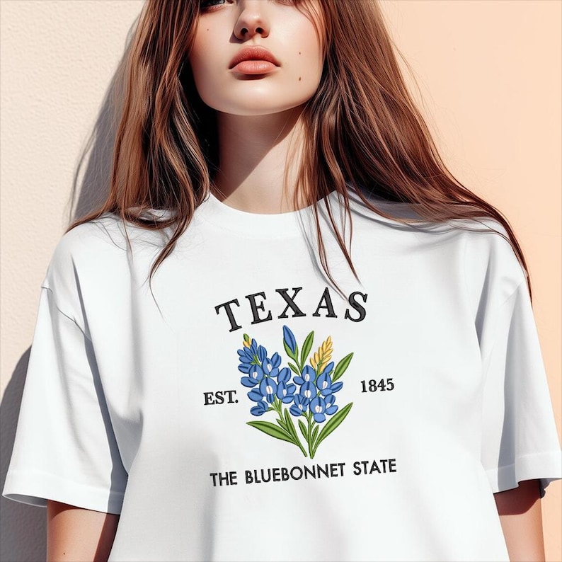 May include: White t-shirt with the word "TEXAS" above a bluebonnet flower design. The shirt also includes the text "EST. 1845" and "THE BLUEBONNET STATE".