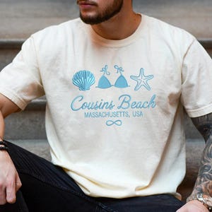 Puede incluir: Camiseta color crema con un dise&ntilde;o azul claro de una concha, un top de bikini y una estrella de mar. El texto "Cousins Beach Massachusetts, USA" est&aacute; impreso debajo, junto con un s&iacute;mbolo de infinito.