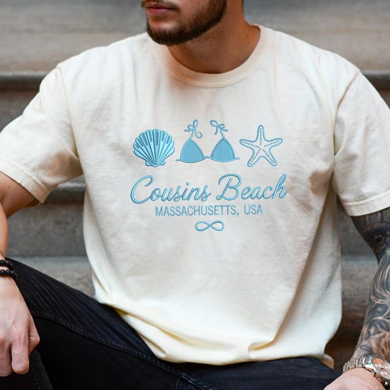 Puede incluir: Camiseta color crema con un dise&ntilde;o azul claro de una concha, un top de bikini y una estrella de mar. El texto "Cousins Beach Massachusetts, USA" est&aacute; impreso debajo, junto con un s&iacute;mbolo de infinito.