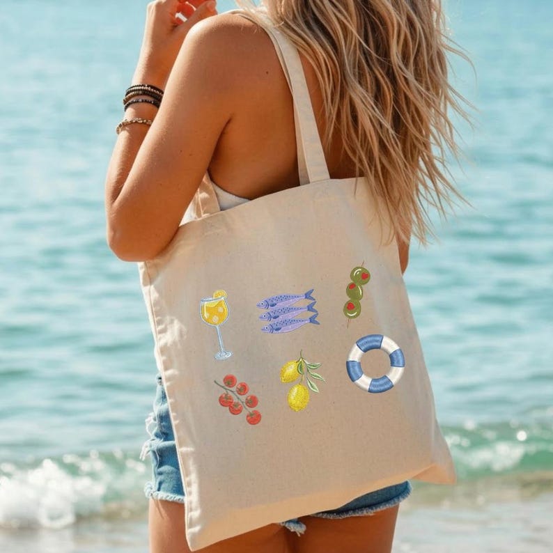 K&ouml;nnte beinhalten: Beige Tragetasche mit farbenfrohen Illustrationen eines Cocktails, Sardinen, Oliven, einer Schwimmweste, Tomaten und Zitronen. Die Tasche wird an einem sonnigen Tag am Meer getragen.