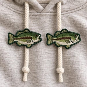 May include: A light gray heathered hoodie featuring two thick, cream-colored braided drawstrings. Each drawstring is adorned with an embroidered patch of a green and brown largemouth bass fish, positioned symmetrically on the front of the garment.