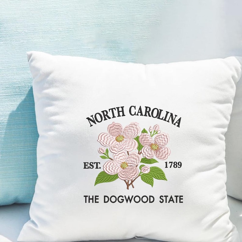 May include: White square pillow with an embroidered floral design and text. The design features pink dogwood flowers, green leaves, and the words "NORTH CAROLINA", "EST. 1789", and "THE DOGWOOD STATE" in black.
