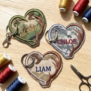 May include: Three heart-shaped fabric keychains with camouflage patterns, each featuring an embroidered deer antler design and a personalized name: Jackson, Chloe, or Liam. Each keychain includes a metal split ring and a lobster clasp.