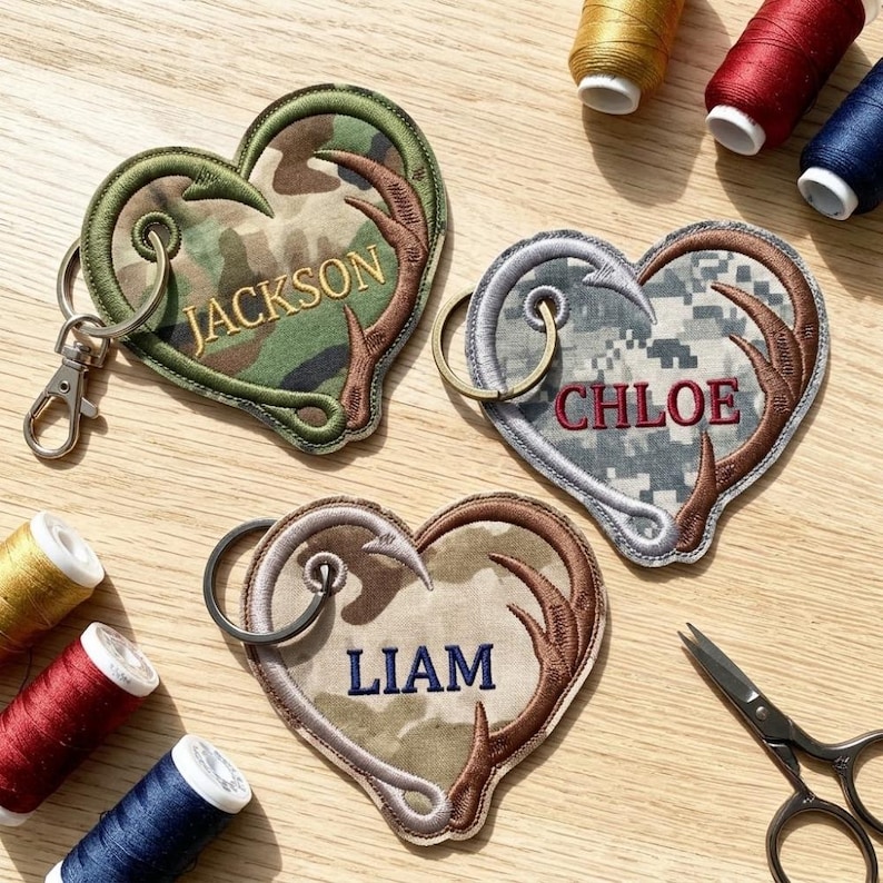 K&ouml;nnte beinhalten: Drei herzf&ouml;rmige Schl&uuml;sselanh&auml;nger aus Stoff mit Tarnmustern, jeweils mit gesticktem Hirschgeweih-Design und einem personalisierten Namen: Jackson, Chloe oder Liam. Jeder Schl&uuml;sselanh&auml;nger enth&auml;lt einen Metallring und einen Karabiner.