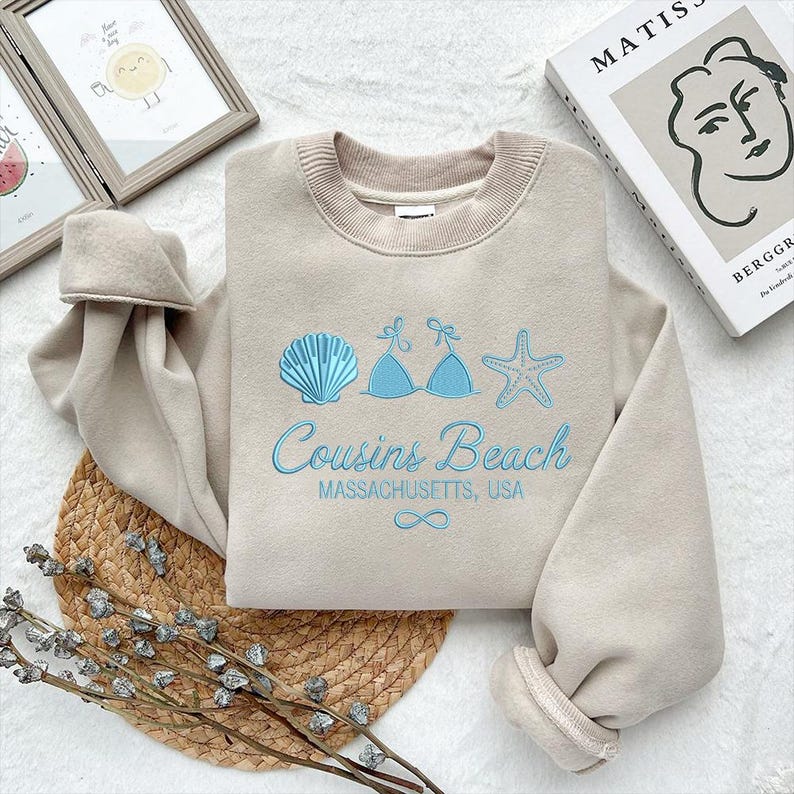 Puede incluir: Sudadera beige con bordados azules de concha, top de bikini y dise&ntilde;os de estrella de mar. El texto "Cousins Beach Massachusetts, USA" est&aacute; bordado en azul. La sudadera est&aacute; doblada sobre una estera tejida.