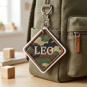 Op de afbeelding: Een ruitvormige tassenhanger van stof met een camouflagepatroon in tinten groen, bruin en zwart. De naam LEO is in beige hoofdletters in het midden geborduurd. De hanger heeft een witte rand en een zilverkleurige metalen karabijnhaak.