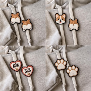 Op de afbeelding: Een set van vier paar geborduurde trekkoordbedels bevestigd aan beige hoodiekoorden. De ontwerpen omvatten Corgi-achterwerken, een Corgi-gezicht, hartvormige patches met de tekst DOG MOM en een pootafdruk, en beige pootafdrukvormen.