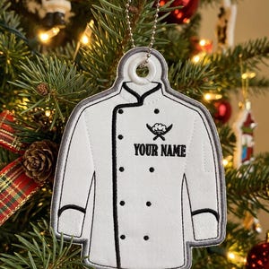 May include: A white chef coat ornament with black trim and buttons, featuring an embroidered chef hat and crossed knives icon above the text YOUR NAME. The ornament is suspended by a silver ball chain and has a grey stitched border.
