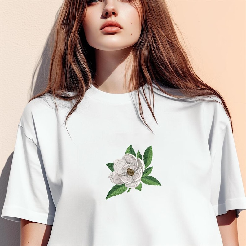 May include: A white short-sleeved t-shirt featuring a centered embroidered design of a single white magnolia flower with a textured brown center and several green leaves. The garment has a crew neckline and a plain, solid white fabric construction.