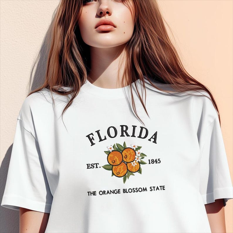 K&ouml;nnte beinhalten: Wei&szlig;es T-Shirt mit dem Wort "FLORIDA" in schwarzer Schrift &uuml;ber einem Orangenbl&uuml;ten-Design. Das Design umfasst orangefarbene Fr&uuml;chte, gr&uuml;ne Bl&auml;tter und wei&szlig;e Bl&uuml;ten. Der Text "EST. 1845" und "THE ORANGE BLOSSOM STATE" sind ebenfalls aufgedruckt.