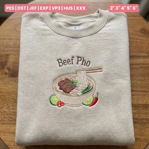 May include: A beige sweatshirt with an embroidered design of a bowl of Beef Pho. The design includes noodles, beef, lime, and chili peppers. The text "Beef Pho" is embroidered above the bowl. The sweatshirt is folded on a wooden surface.