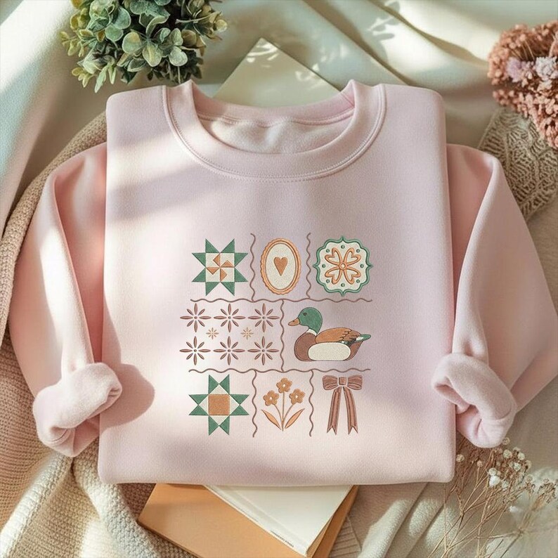 May include: A light pink sweatshirt with a crew neck and long sleeves. The front features a patchwork design with a duck illustration, floral and geometric patterns. The design is in shades of green, brown, and pink.