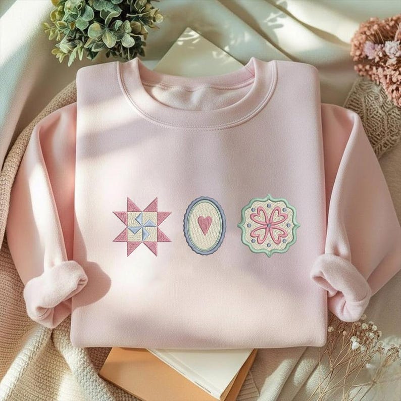 May include: A light pink sweatshirt with three embroidered patches. The patches include a starburst, an oval with a heart, and a floral design. The sweatshirt has a crew neck and long sleeves with cuffed wrists.