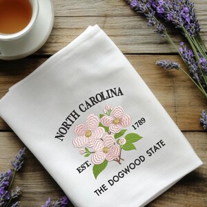 May include: White kitchen towel embroidered with pink dogwood flowers, green leaves, and the text "NORTH CAROLINA" and "THE DOGWOOD STATE". Includes the year "1789".