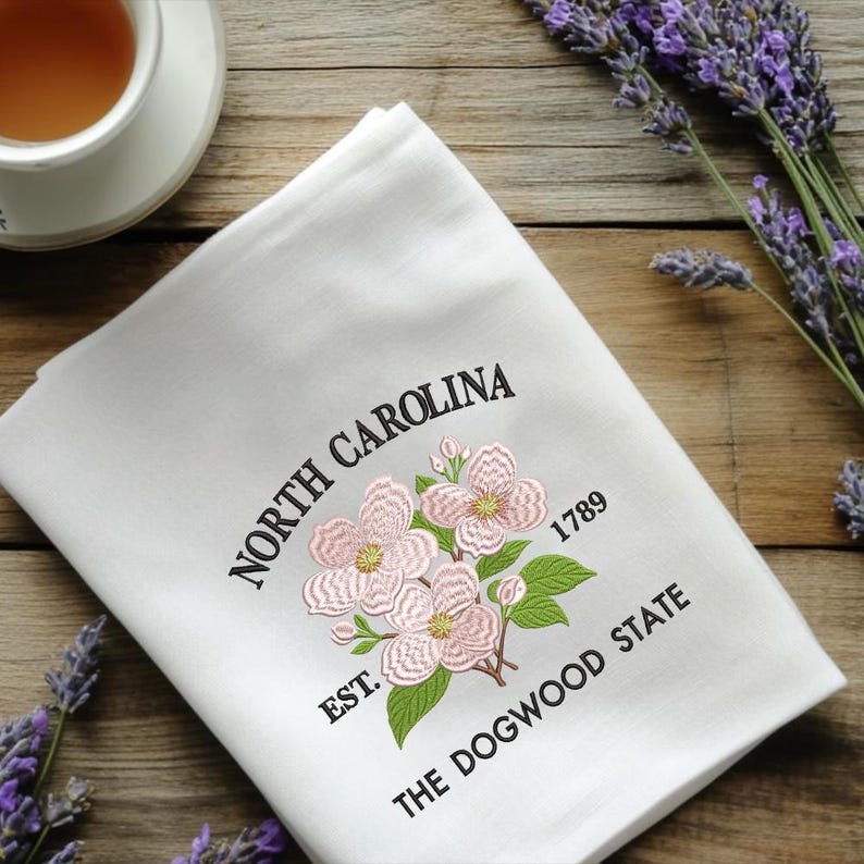 May include: White kitchen towel embroidered with pink dogwood flowers, green leaves, and the text "NORTH CAROLINA" and "THE DOGWOOD STATE". Includes the year "1789".