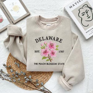 May include: Beige sweatshirt featuring the word "DELAWARE" in black script, embroidered pink flowers, and the text "EST. 1787 THE PEACH BLOSSOM STATE". The sweatshirt has a crew neck and long sleeves.